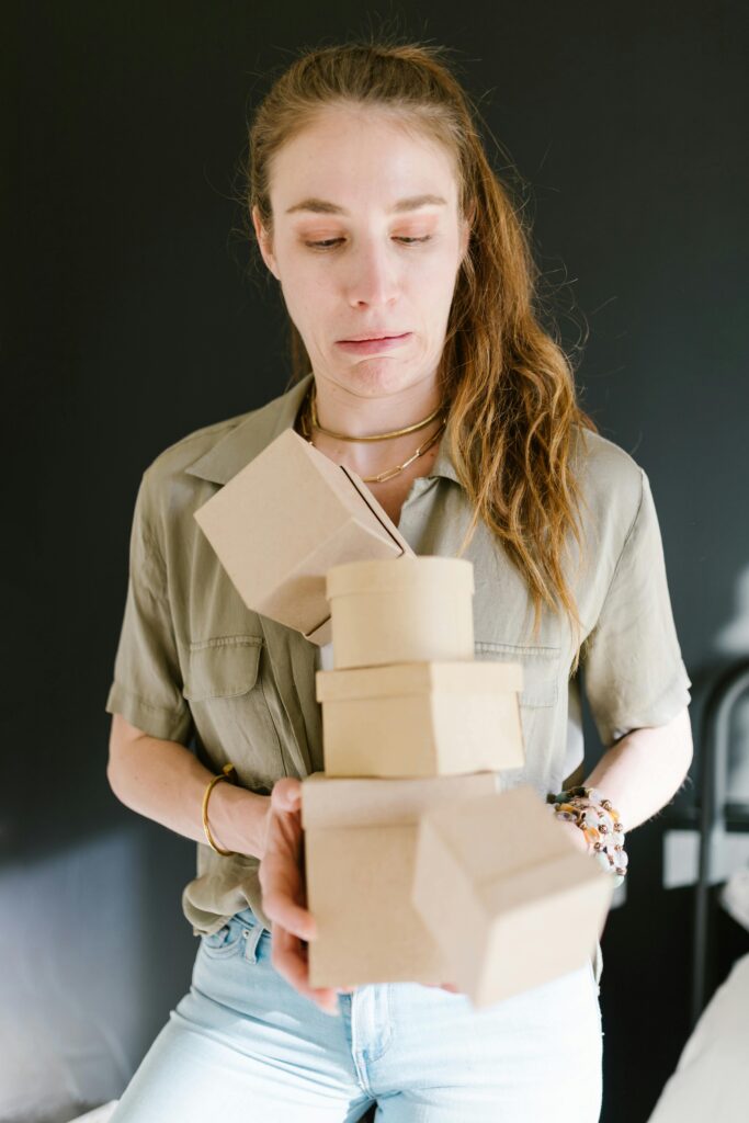 A woman in a green shirt struggles to balance a stack of cardboard boxes indoors.